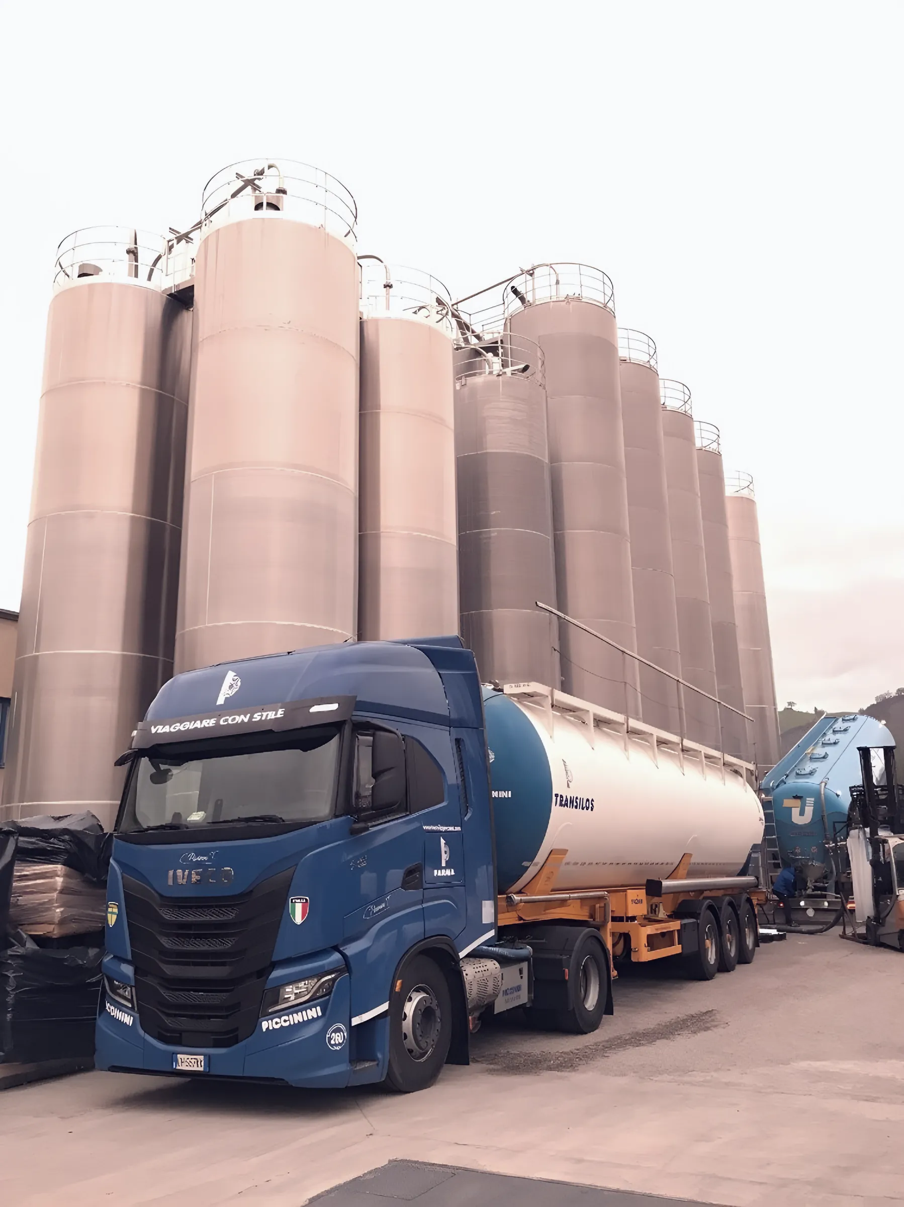 Blue Piccinini Iveco tractor with silo trailer beneath a row of vertical silos at a plant