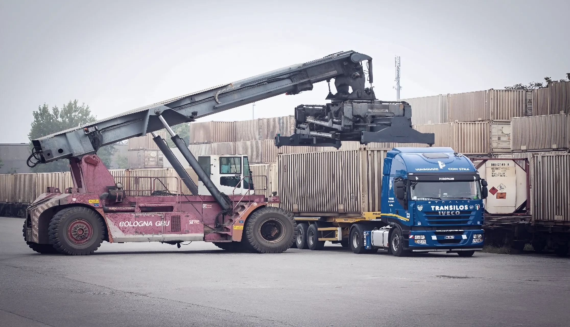 Red reach stacker lifting a container above a Transilos Piccinini Iveco tractor