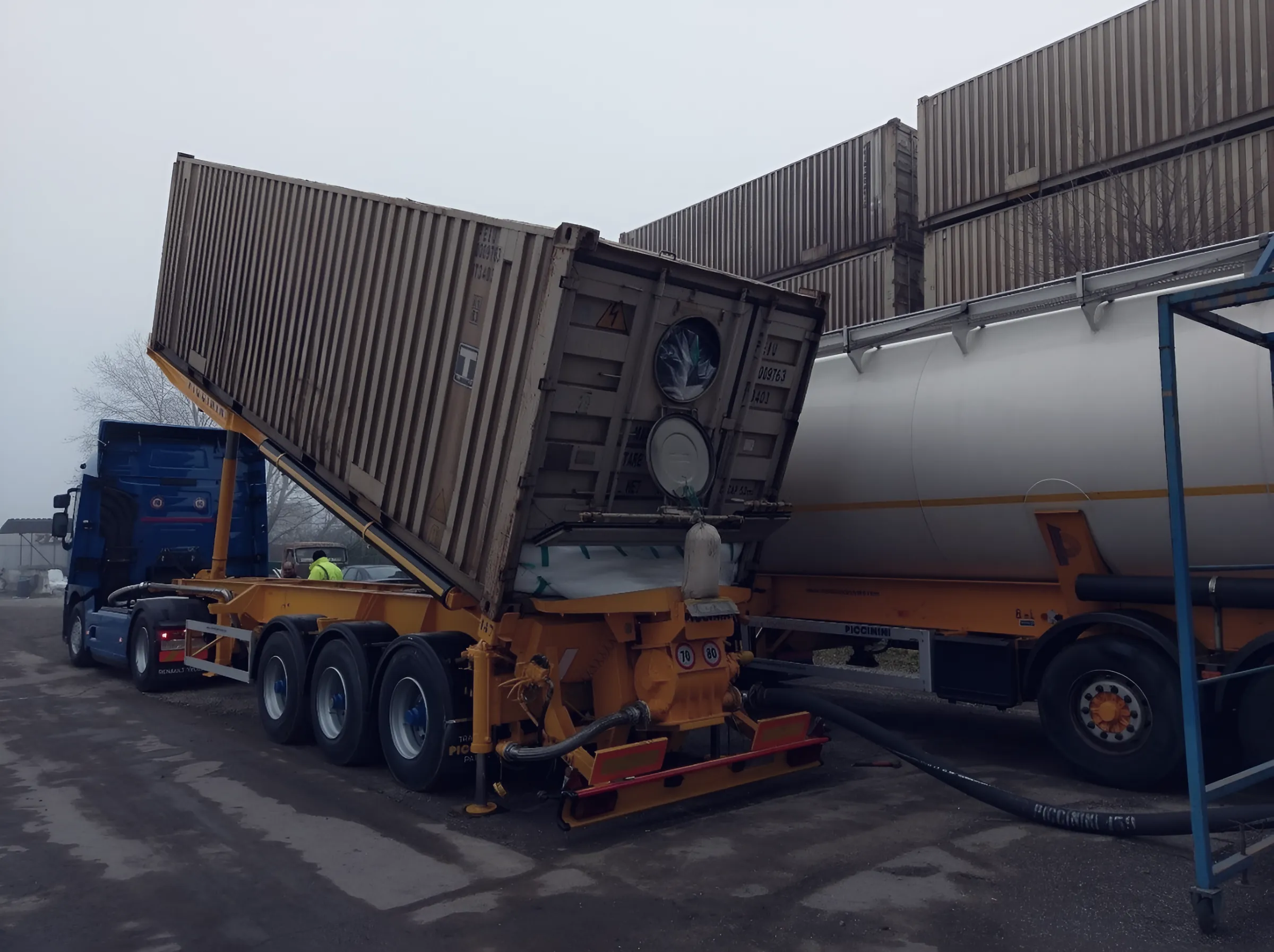 Container tipped on a yellow chassis connected by hose to a silo trailer