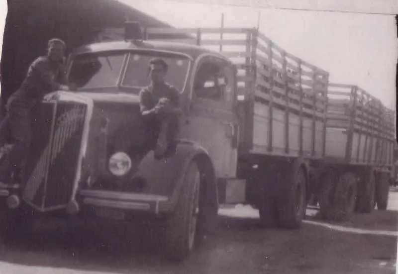 Two drivers sitting on the bonnet of a lorry with wooden sides and trailer