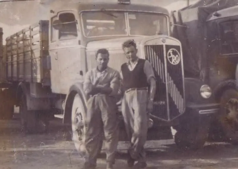 Two drivers in front of the grille of a vintage lorry with wooden sides