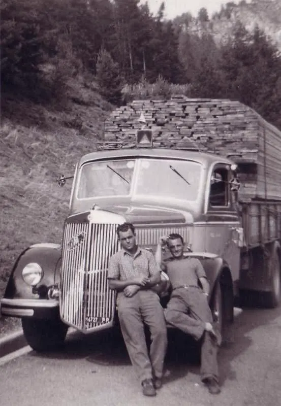 Two drivers sitting on the bumper of a timber-laden lorry on a mountain road